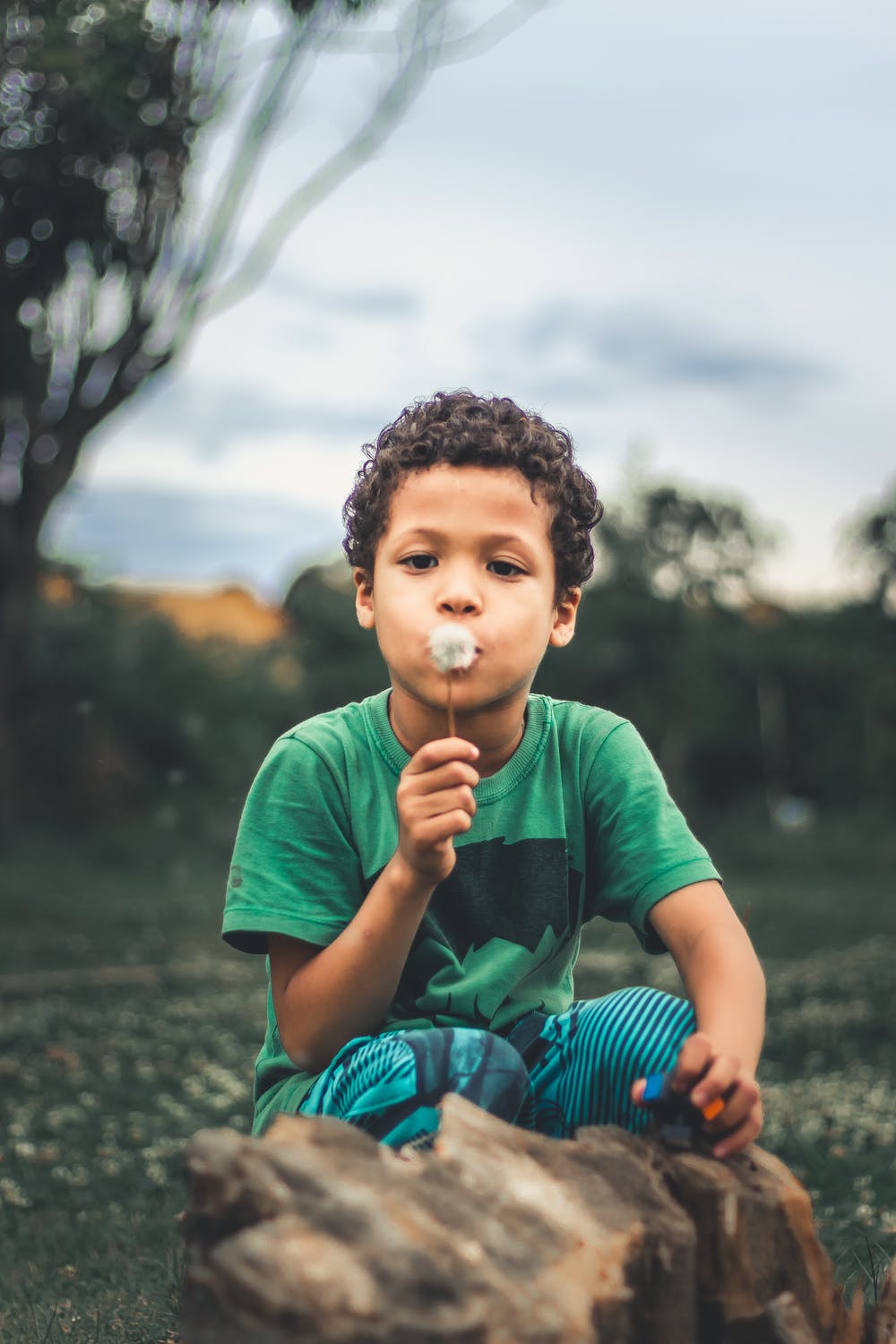 A child wearing a green shirt is blowing on a dandelion in a grassy outdoor setting.